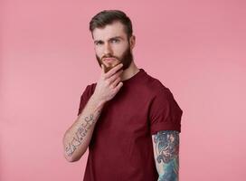 portret van jong aantrekkelijk denken getatoeëerd rood gebaard Mens in rood t-shirt, looks Bij de camera en accenten de kin, staat over- roze achtergrond. foto