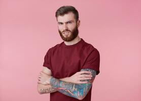 portret van jong getatoeëerd walgde rood gebaard Mens in blanco t-shirt, staat met gekruiste armen over- roze achtergrond, fronsen en looks Bij de camera. foto