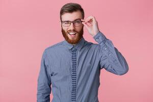 portret van jong glimlachen aantrekkelijk gebaard Mens in een gestreept shirt, houdt bril en knipoogt geïsoleerd over- roze achtergrond. foto