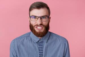 dichtbij omhoog van niet tevreden boos jong knap rood gebaard Mens met bril en een gestreept shirt, staat over- roze achtergrond, uitbarstingen zijn tanden agressief en looks in de camera, looks gek. foto