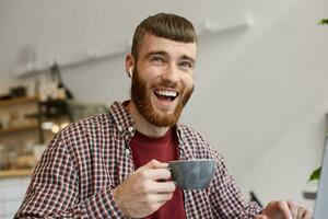 portret van een jong knap roodbaard vent breed glimlachen en lacht Bij een grappig grap , genieten van heerlijk gebrouwen koffie, vervelend in eenvoudig kleren. foto