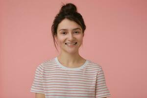 tiener- meisje, gelukkig op zoek vrouw met brunette haar- en broodje. vervelend t-shirt met rood stroken en groot glimlach. emotioneel concept. aan het kijken Bij de camera geïsoleerd over- pastel roze achtergrond foto