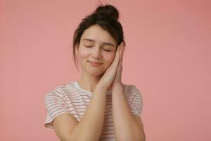 tiener- meisje, gelukkig op zoek vrouw met brunette haar- en broodje. vervelend t-shirt met rood stroken en doen alsof ze is slapen, dromen. emotioneel concept. staan geïsoleerd over- pastel roze achtergrond foto