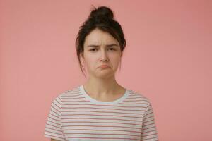 jong dame, ontevreden vrouw met brunette haar- en broodje. vervelend t-shirt met rood stroken en op zoek beledigd. emotioneel concept. aan het kijken Bij de camera geïsoleerd over- pastel roze achtergrond foto