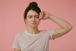 portret van aantrekkelijk, van streek meisje met brunette haar- en broodje. vervelend t-shirt met rood stroken en wrijven een oog. emotioneel concept. staan geïsoleerd over- pastel roze achtergrond foto