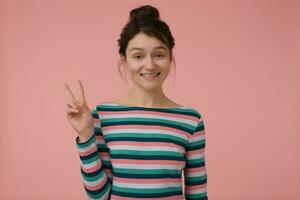 tiener- meisje, gelukkig op zoek vrouw met brunette haar- en broodje. vervelend gestreept blouse en tonen vrede teken, glimlach. emotioneel concept. aan het kijken Bij de camera geïsoleerd over- pastel roze achtergrond foto