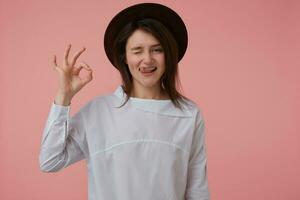 tiener- meisje, gelukkig op zoek vrouw met lang brunette haar. vervelend wit blouse en zwart hoed. tonen Oke teken. emotioneel concept. aan het kijken en knipoog Bij de camera geïsoleerd over- pastel roze achtergrond foto