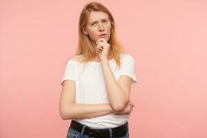 nadenkend jong aantrekkelijk roodharige vrouw met gewoontjes kapsel houden verheven hand- Aan haar kin en loensen ogen terwijl op zoek bedachtzaam Bij camera, poseren over- roze achtergrond foto