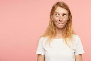 verbijsterd jong lief groene ogen roodharige vrouw met gewoontjes kapsel bijten haar lippen terwijl op zoek omhoog, poseren over- roze achtergrond in wit eenvoudig t-shirt foto