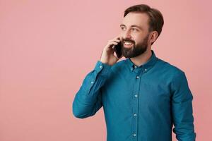 blij aantrekkelijk gebaard Mens, op zoek weg en lachend, vervelend een denim shirt, pratend Aan de telefoon. geïsoleerd over- roze achtergrond. foto