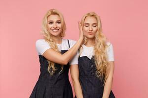 gelukkig jong bruine ogen lang haren vrouw glimlachen breed terwijl op zoek Bij camera en aan het bedekken oren van haar mooi blond zus terwijl maken haar verrassing, geïsoleerd over- roze achtergrond foto