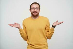 studio foto van jong knap brunette gebaard mannetje schouderophalend met verheven palmen en glimlachen een beetje Bij camera, vervelend mosterd trui en polshorloge over- wit achtergrond