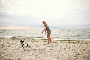 horizontaal buitenshuis schot van mooi jong vrouw met blond haar- wandelen langs strand Aan bewolkt met haar hond, vervelend zomer jurk en rietje hoed foto