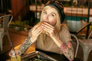 charmant getatoeëerd blond vrouw met lang haar- op zoek Bij camera terwijl proberen Hamburger in stad cafe gedurende lunch pauze, vervelend beige t-shirt en leer zwart baret foto