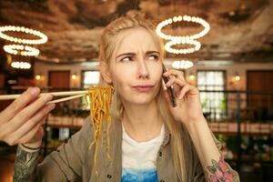 portret van echt op zoek getatoeëerd blond vrouw met gewoontjes kapsel Holding noedels met houten stokjes terwijl pratend Aan telefoon, vervelend beige overhemd en wit t-shirt foto