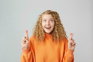 charmant positief meisje vriendelijke groet geloven in haar geluk. schot van jong gekruld gelukkig glimlachen geïnspireerd blond vrouw houden haar vingers gekruiste en ogen op zoek omhoog terwijl maken een wens foto