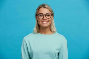 portret van gelukkig charmant vrolijk blond jong meisje in bril, glimlacht, op zoek gelukkig en blij, levendig, met natuurlijk verzinnen, gebruind, gekleed in gewoontjes licht blauw lang mouw t-shirt, blauw achtergrond foto