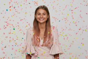 detailopname van vrolijk aantrekkelijk jong vrouw met lang geverfd pastel roze haar- draagt polka punt roze jurk hebben pret en vieren verjaardag geïsoleerd over- wit achtergrond met confetti foto