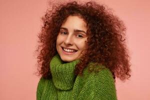 portret van aantrekkelijk, volwassen meisje met gember gekruld haar. vervelend groen coltrui trui en lachend. aan het kijken flirterig Bij de camera geïsoleerd, detailopname over- pastel roze achtergrond foto