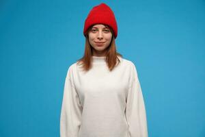 tiener- meisje, koel op zoek vrouw met brunette haar. vervelend wit trui en rood hoed. mensen en emotioneel concept. aan het kijken Bij de camera en glimlach, geïsoleerd over- blauw achtergrond foto