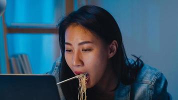 freelance slimme zakenvrouwen die instant noedels eten, moe zijn terwijl ze 's nachts thuis op een laptop in de woonkamer werken. gelukkig jong aziatisch meisje zittend op bureau werk overuren, geniet van ontspannen tijd.