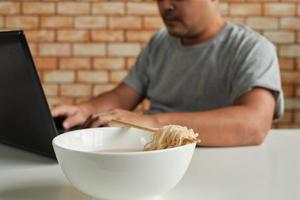 Thaise mannelijke werknemer druk aan het werk met laptop, gebruik eetstokjes om haastig instant noedels te eten tijdens de kantoorlunch, want snel, smakelijk en goedkoop. na verloop van tijd Aziatisch fastfood, ongezonde levensstijl. foto