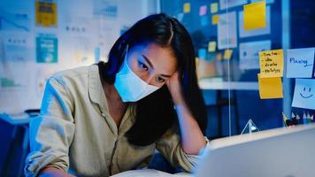 freelance azië vrouwen dragen gezichtsmasker met behulp van laptop hard werken op nieuw normaal kantoor. 's nachts thuiswerken, zelfisolatie, sociale afstand, quarantaine voor preventie van het coronavirus. foto