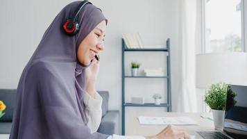 azië moslim dame draagt hoofdtelefoon horloge webinar luister online cursus communiceer via een videoconferentiegesprek thuis. thuiswerken op afstand, sociale afstand, quarantaine voor preventie van het coronavirus.