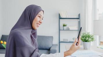 jonge azië moslim zakenvrouw met behulp van slimme telefoon praten met vriend door videochat brainstorm online vergadering terwijl op afstand werken vanuit huis in de woonkamer. sociale afstand, quarantaine voor het coronavirus. foto