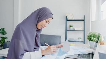 mooie azië moslimdame in hoofddoek vrijetijdskleding met behulp van laptop in de woonkamer in huis. thuiswerken op afstand, nieuwe normale levensstijl, sociale afstand, quarantaine voor preventie van het coronavirus.