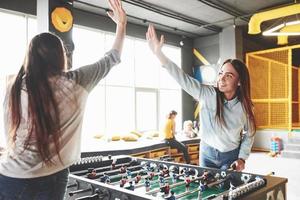 twee mooie tweelingmeisjes spelen tafelvoetbal en hebben plezier. een van de zussen houdt een speelgoedbal in zijn hand en laat de tong zien foto