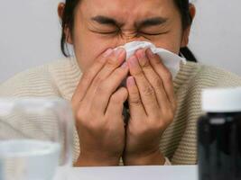 ongezond Aziatisch vrouw in trui blazen neus- in beschikbaar zakdoek. allergie naar stof, dier huidschilfers, koud, of virale infectie. gezond en geneeskunde concept. foto