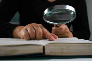 detailopname van een vrouw op zoek door een vergroten glas Bij een leerboek. vergroten glas in hand- en Open boek Aan tafel. onderwijs en Onderzoek concept. foto