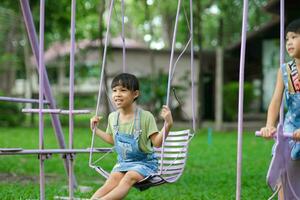 twee schattig weinig zussen hebben pret spelen in de speelplaats gedurende zomer. schattig weinig meisje swinging in de speelplaats met een glimlach en gelach. actief zomer vrije tijd voor kinderen. foto