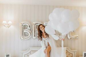 jong gekruld meisje met lang haar- in luxueus kamer staand Aan bed in wit Open jurk Holding veel gel opblaasbaar ballen in handen en voorbereidingen treffen voor bruiloft ceremonie foto
