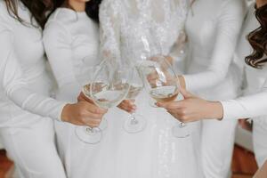 jong bruidsmeisjes in wit zijde jurken drinken Champagne in de bruid kamer. mooi Dames vieren vrijgezellin partij staand in kamer en met Champagne. detailopname foto van handen met bril