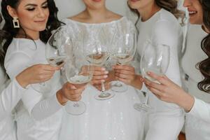 jong bruidsmeisjes in wit zijde jurken drinken Champagne in de bruid kamer. mooi Dames vieren vrijgezellin partij staand in kamer en met Champagne. detailopname foto van handen met bril