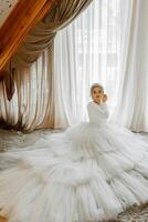 portret van een mooi bruid in een wit bruiloft jurk met een lang trein in een hotel kamer. de bruid is zittend Aan de verdieping tegen een groot venster foto