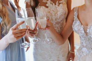 jong bruidsmeisjes drinken Champagne in de bruid kamer. mooi Dames vieren vrijgezellin partij staand in kamer en met Champagne. detailopname foto van handen in bril