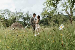 een paar in bruiloft kleren met een boeket van bloemen en groen in hun handen Aan de achtergrond van een veld- met madeliefjes foto