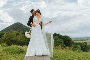 elegant bruid en bruidegom knuffel en kus tegen de achtergrond van zomer bergen. de concept van een rustiek bruiloft in de bergen, gelukkig Boheems pasgetrouwden. foto