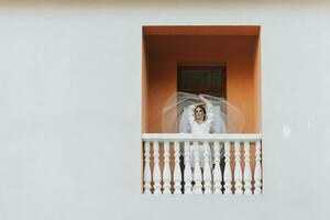 de bruid in een wit gewaad, staand Aan de balkon, gooit omhoog de sluier. voorkant foto. vrij ruimte foto