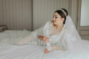 een mooi meisje met een bruiloft kapsel en een tiara Aan haar hoofd in een transparant gewaad, aan het liegen Aan een bed in een hotel met een Koninklijk interieur. lang sluier, natuurlijk licht, portret foto