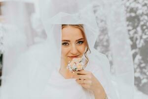 winter bruid, portret van een mooi bruid in een wit poncho en bruiloft jurk in een winter park. de bruid onder een sluier met de bruidegom bloem in zijn handen foto