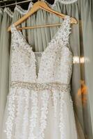 detailopname van een handgemaakt bruiloft jurk Aan een houten hanger met geborduurd bloemen. in de buurt de jurk met Open schouders zonder mouwen foto