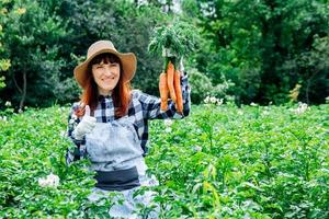 vrouw boer houdt een bos wortelen op de achtergrond moestuin stro hoed en omringd door de vele planten in haar moestuin