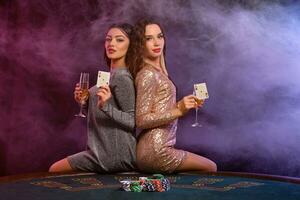 twee meisjes zijn spelen poker Bij casino, Holding bril van Champagne en kaarten, zittend Aan tafel met chips Aan het. zwart, rook achtergrond. detailopname. foto