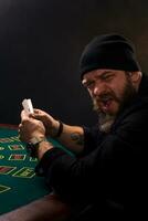 echt gebaard Mens zittend Bij poker tafel en Holding kaarten geïsoleerd Aan zwart. kopiëren ruimte foto