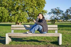 portret van jong Aziatisch vrouw zittend Aan bank met smartphone. schattig meisje gebruik makend van mobiel telefoon, genieten van wezen buitenshuis foto