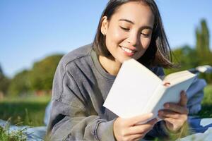 portret van mooi glimlachen Aziatisch meisje, lezing in park, aan het liegen Aan gras met favoriete boek. vrije tijd en mensen concept foto